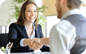 A smiling businesswoman in a black blazer shaking hands with a man in a professional setting, suggesting a successful meeting or agreement.